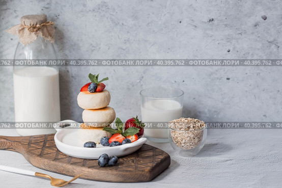 Cottage cheese pancakes, Russian cheesecakes with fresh blueberries, strawberries and mint on a stylish wooden board on a gray concrete table. Natural products. Healthy and delicious breakfast. Стоковое фото, фотограф Photojuli / Фотобанк Лори