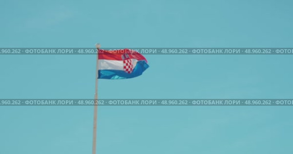 Croatian national flag waving proudly on a pole against a bright clear blue sky. Стоковое видео, видеограф Igor Tichonow / Фотобанк Лори