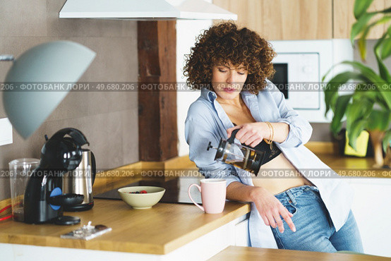 Curly haired female in unbuttoned shirt and black bra pouring hot tea into mug while leaning on electric stove. Woman pouring tea from french press into cup in modern kitchen. Стоковое фото, агентство Ingram Publishing / Фотобанк Лори