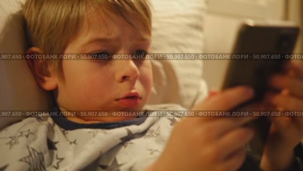 Cute kid boy in pajamas lying in bed watching smartphone, smiling. Children's screen time. Child plays games using phone at the evening or night. Social media addiction. Childhood, modern technology. Стоковое видео, видеограф Марина Демидюк / Фотобанк Лори