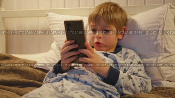 Cute kid boy in pajamas lying in bed watching smartphone, smiling. Children's screen time. Child plays games using phone at the evening or night. Social media addiction. Childhood, modern technology. Стоковое видео, видеограф Марина Демидюк / Фотобанк Лори