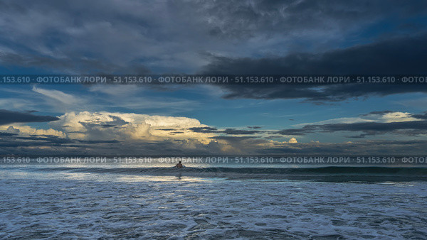 Dawn over the ocean. The waves roll towards the shore, spreading, foaming. The silhouette of a man in the water. The clouds in the blue sky are highlighted in gold. Early morning. Malaysia. Borneo. Стоковое фото, фотограф Вера Ивановна Кубарева / Фотобанк Лори