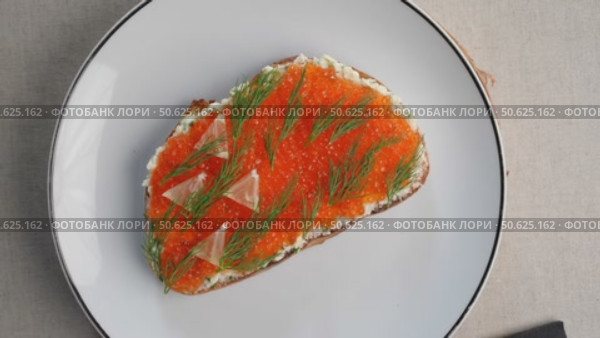 Delicious toast with cream cheese, red caviar and lemon slices, a traditional Eastern European appetizer, is presented on a white plate with black cutlery. Стоковое видео, видеограф Данил Руденко / Фотобанк Лори