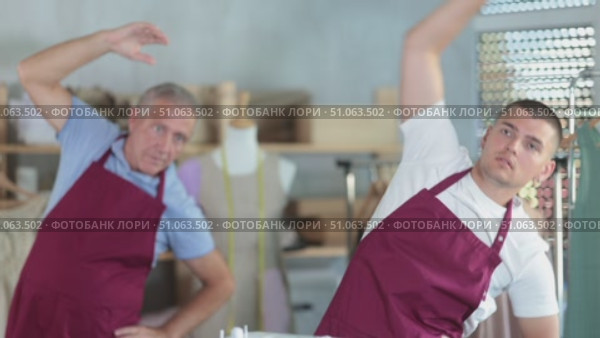 Elderly and young male tailors doing warm-up exercises while working in sewing workshop. Стоковое видео, видеограф Яков Филимонов / Фотобанк Лори