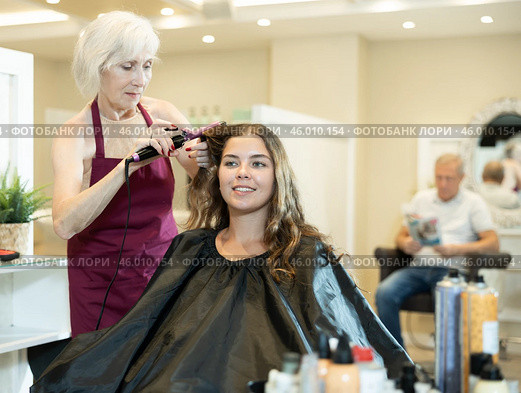 Elderly female hairstylist using curling wands to add volume for hair of young girl. Стоковое фото, фотограф Яков Филимонов / Фотобанк Лори