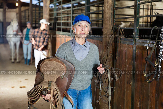 Elderly female rancher walking in stable with saddle in hands. Стоковое фото, фотограф Яков Филимонов / Фотобанк Лори