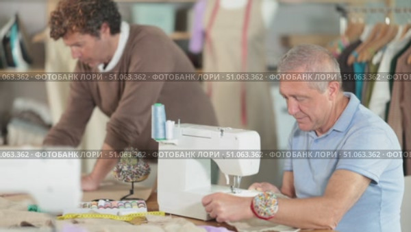 Elderly man tailor sews on machine adult man assistant drawing pattern on paper in workshop. Стоковое видео, видеограф Яков Филимонов / Фотобанк Лори