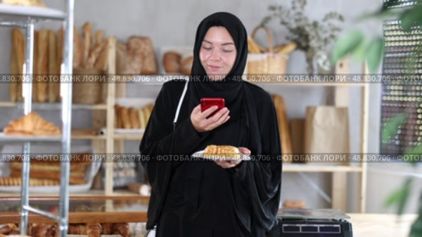 European Muslim girl customer holds pastry on disposable plate and recognizes it using app. Стоковое видео, видеограф Яков Филимонов / Фотобанк Лори