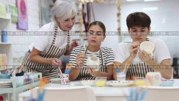 Experienced mature female ceramicist teaching young enthusiastic couple, girl and guy, art of ceramic glazing and painting in pottery studio. Стоковое видео, видеограф Яков Филимонов / Фотобанк Лори