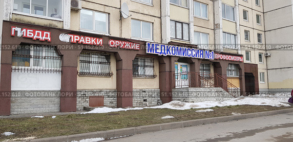 Facade of a multi-story residential building with commercial premises located on the ground floor. The building has bright signs of organizations and services: "Traffic Police", "Certificates", "Weapons", "Medical Commission No. 1", "Medical Examinations". A porch with stairs and railings leading to the entrance is visible. The windows are protected by metal bars. The lower part of the facade is made of gray brick and stone cladding, damage and cracks are visible on it. There are still remnants of snow on the street, and patches of green grass are visible near the sidewalk. The atmosphere is typical of the urban environment of Russia with the characteristic architecture of panel houses and a variety of commercial advertisements. (2019 год). Редакционное фото, фотограф Анна Кривицкая / Фотобанк Лори