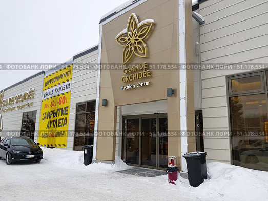 Facade of the Grande Orchidee fashion center in winter. In the foreground you can see the entrance with large glass doors and a large golden emblem in the form of an orchid, as well as the name of the center. On the left there is a large yellow banner with the inscription about the outlet closing and discounts from 30% to 80%, the text is duplicated in several languages ​​(Russian, Finnish). There is a black car in front of the building, snow lies on the ground, trash cans and a few snowdrifts are visible near the entrance. The atmosphere of the photo conveys a winter day at the entrance to a fashionable shopping center during a sale and liquidation of goods. (2019 год). Редакционное фото, фотограф Анна Кривицкая / Фотобанк Лори