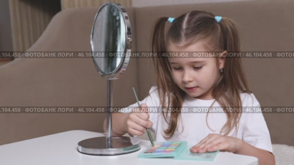 Fashion little girl in a white T-shirt is sitting at the dressing table in front of the mirror and applying makeup with eye shadow. Advertising of children's cosmetics concept. Footage for kids blog. Стоковое видео, видеограф Ирина Михайличенко / Фотобанк Лори