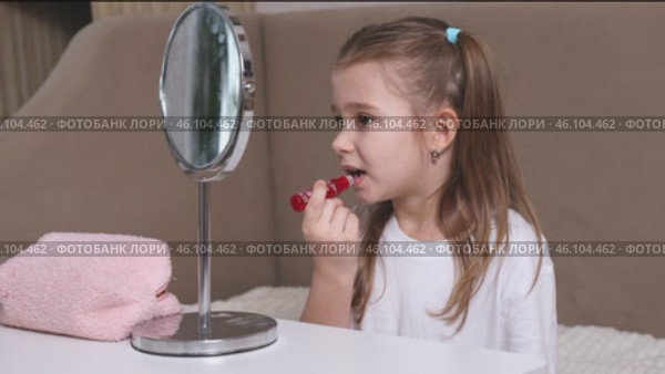 Fashion little girl in a white T-shirt is sitting at the dressing table in front of the mirror and applying makeup with lipstick. Advertising of children's cosmetics concept. Footage for kids blog. Стоковое видео, видеограф Ирина Михайличенко / Фотобанк Лори