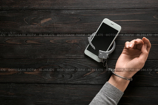Female hand in handcuffs with mobile phone on wooden background. Concept of addiction. Стоковое фото, фотограф Pixel_shot / Фотобанк Лори