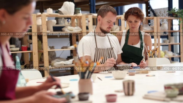Female teacher and mentor helps students learn how to make patterns on ceramic plates using paint and brush in a pottery workshop. Стоковое видео, видеограф Яков Филимонов / Фотобанк Лори