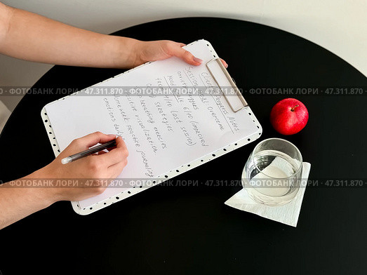 Female writing on clipboard with notes next to apple and glass on round black table. Стоковое фото, фотограф Lamina Akulova / Фотобанк Лори