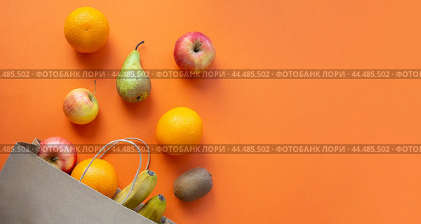 Flat lay of fruits, including an apple, pear, bananas, oranges and a kiwi, arranged around a brown paper bag on an orange background. Стоковое фото, фотограф Ольга Петрова-Апостолова / Фотобанк Лори