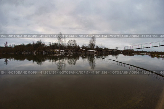 Фотография заснеженного подвесного деревянного моста над рекой Десна в сельской местности России. Мрачный день. Белый и серый цвета. Концепции туризма и образа жизни. Линза «рыбий глаз». Стоковое фото, фотограф YuryKara / Фотобанк Лори