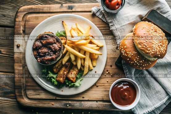 French fries, burger, pork ribs on a stylish board on a wooden background. Fried potatoes. The concept of fast food, junk food and fast food preparation. Стоковое AI-изображение, AI-художник Photojuli / Фотобанк Лори