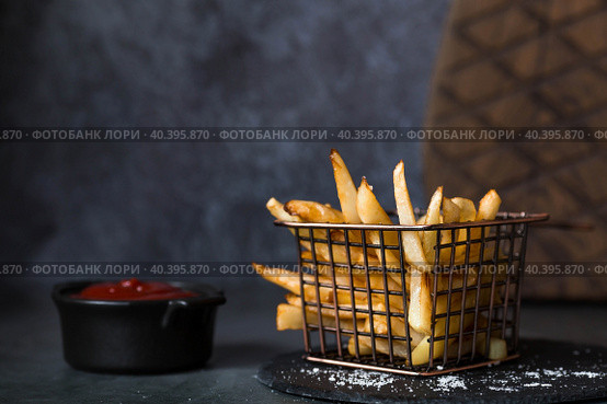 French fries in a metal basket with salt and ketchup on a stylish board on a dark background. Fried potatoes. The concept of fast food and junk food. Стоковое фото, фотограф Photojuli / Фотобанк Лори
