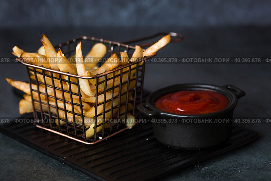 French fries in a metal basket with salt and ketchup on a stylish board on a dark background. Fried potatoes. The concept of fast food and junk food. Стоковое фото, фотограф Photojuli / Фотобанк Лори