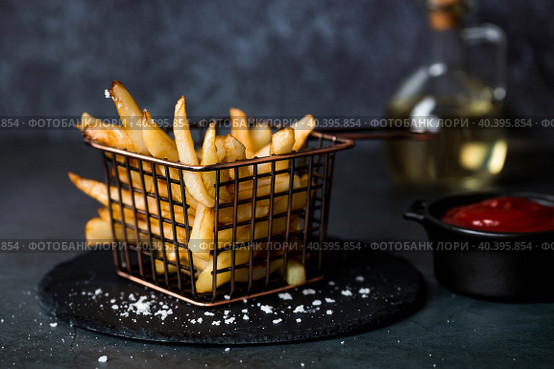 French fries in a metal basket with salt, ketchup and sunflower oil on a stylish board on a dark background. Fried potatoes. The concept of fast food and junk food. Стоковое фото, фотограф Photojuli / Фотобанк Лори