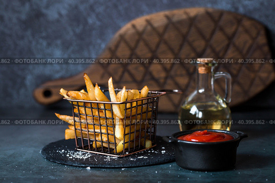 French fries in a metal basket with salt, ketchup and sunflower oil on a stylish board on a dark background. Fried potatoes. The concept of fast food and junk food. Стоковое фото, фотограф Photojuli / Фотобанк Лори