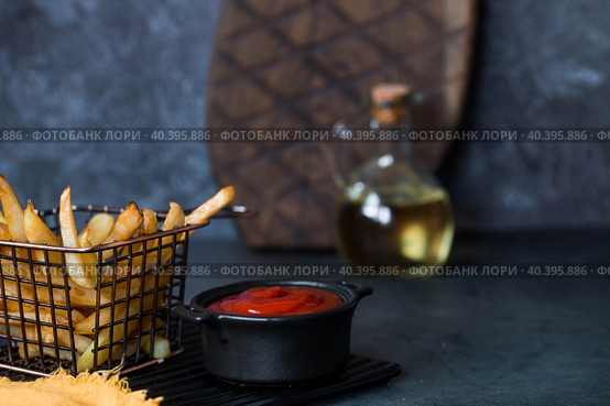 French fries in a metal basket with salt, ketchup and sunflower oil on a stylish board on a dark background. Fried potatoes. The concept of fast food and junk food. Стоковое фото, фотограф Photojuli / Фотобанк Лори