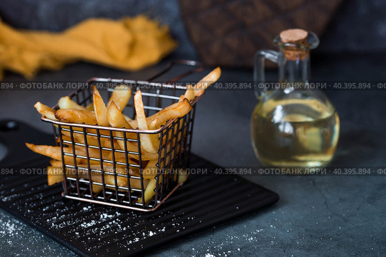 French fries in a metal basket with salt, ketchup and sunflower oil on a stylish board on a dark background. Fried potatoes. The concept of fast food and junk food. Стоковое фото, фотограф Photojuli / Фотобанк Лори