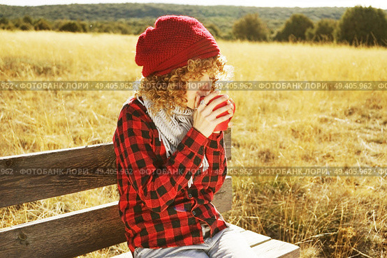 Front portrait of a young woman in red plaid shirt with a wool cap and scarf taking a cup of tea or coffee while she is sunbathing sitting in a wooden bench in a yellow field with backlight from autumn sun. Стоковое фото, агентство Ingram Publishing / Фотобанк Лори