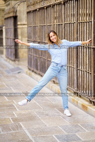 Full body cheerful adult female in casual clothes raising arm and leg and looking at camera with smile, while grasping bar of aged fence on city street. Playful woman grasping old fence. Стоковое фото, агентство Ingram Publishing / Фотобанк Лори