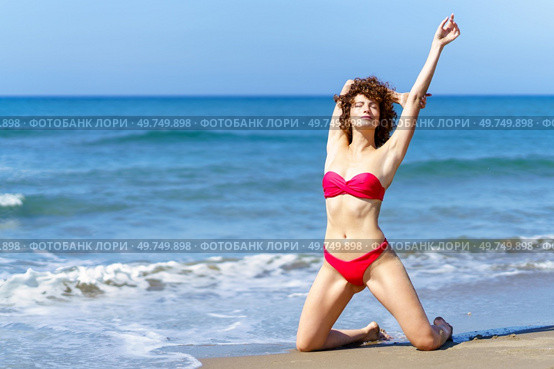 Full body of tender woman with curly hair and in red bikini raising arm while standing on knees on sandy beach of ocean and sunbathing. Sensual woman resting on sandy shoreline. Стоковое фото, агентство Ingram Publishing / Фотобанк Лори