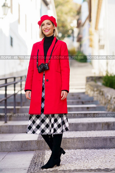 Full length of happy adult female tourist in elegant coat and beret with photo camera on neck smiling and looking away while standing on stone stair in old town. Stylish woman in red coat standing on city street in sunlight. Стоковое фото, агентство Ingram Publishing / Фотобанк Лори