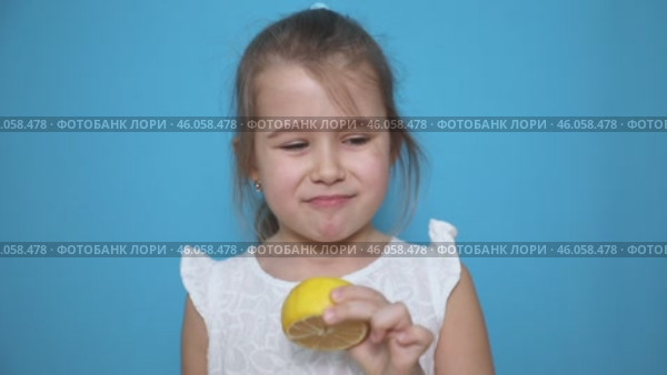 Funny little child girl try to taste fresh lemon, wrinkles and makes a grimace. Sour taste, facial emotions negative, in white dress, isolated blue background, copy space. Стоковое видео, видеограф Ирина Михайличенко / Фотобанк Лори