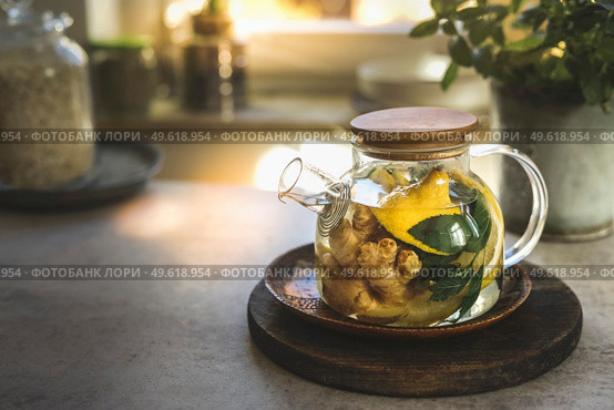 Glass tea pot with mint leaves, lemon and ginger on kitchen table with utensils and window background. Herbal tea with healthy ingredients and vitamin c in cold season. Natural treatment. Front view. Стоковое фото, агентство Ingram Publishing / Фотобанк Лори