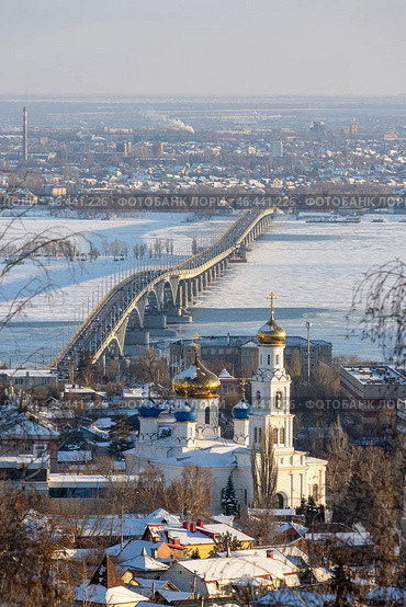 Город Саратов, Саратовская область, Россия. Вид сверху на Храм Сошествия Святого Духа и Саратовский мост через Волгу. Зимний городской пейзаж. На переднем плане красивая белая православная церковь с колокольней. Вдали автомобильный мост, соединяющий город Саратов и город Энгельс (расположенный на противоположном берегу Волгоградского водохранилища). Стоковое фото, фотограф Андрей Степанов / Фотобанк Лори