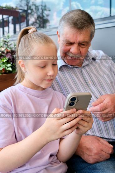 Granddaughter showing smartphone to grandfather. Portrait of a cute girl playing or typing on her smartphone while sitting in a cafe with her grandfather. Стоковое фото, фотограф Терехова Марина / Фотобанк Лори