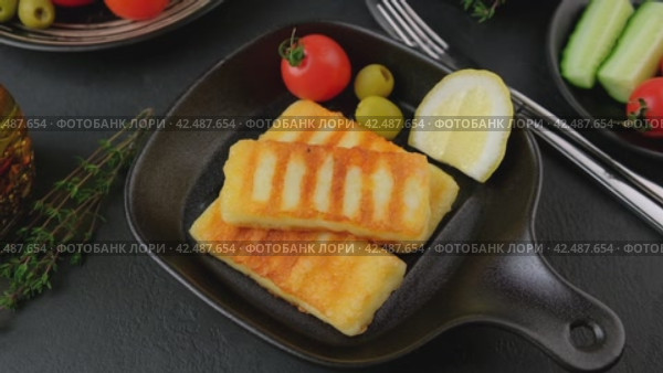 Grilled halloumi cheese in a black pan with olives, tomatoes, cucumbers, thyme and lemon. Traditional cheese of the Eastern Mediterranean. Black background, selective focus, close-up. Slow motion. Стоковое видео, видеограф Наталья Любина / Фотобанк Лори