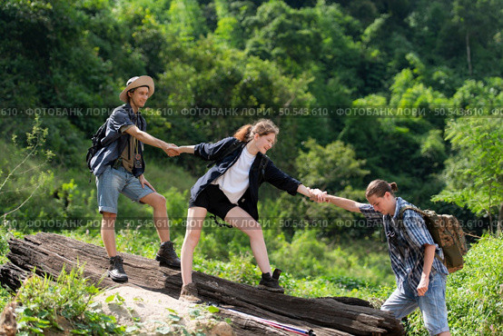 Group of friend on a sunny day in the forest Young group hiker with backpack and trekking poles helping to pull each other up to stand on the log. Стоковое фото, агентство Ingram Publishing / Фотобанк Лори