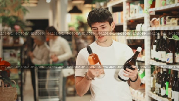 Guy chooses semi-sweet wine while standing near the shelves with alcohol in a hypermarket. European chooses bottles of wine in a store against the background of customers. Стоковое видео, видеограф Яков Филимонов / Фотобанк Лори