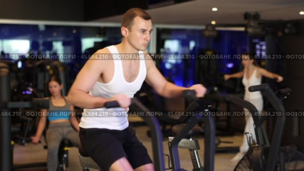 Guy performs exercises with stationary air bicycle in gym. Стоковое видео, видеограф Яков Филимонов / Фотобанк Лори