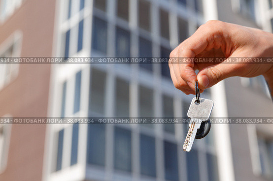 Hand holds the keys to a new apartment on the background of the house. Housewarming and construction. Рука держит ключи от новой квартиры на фоне дома новостройки. Новоселье, ремонт и строительство. Стоковое фото, фотограф Aleksandra Selivanova / Фотобанк Лори