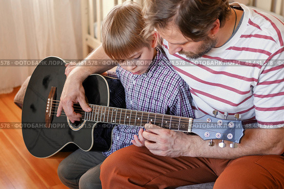 Handsome dad teaches his cute son to play the guitar sitting on sofa in the living room at home. Happy Father's Day. Father teaching his child to play guitar. Kid Learning music skill. Art education. Стоковое фото, фотограф Марина Демидюк / Фотобанк Лори