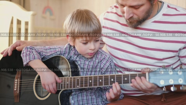 Handsome dad teaches his cute son to play the guitar sitting on sofa in the living room at home. Happy Father's Day. Father teaching his child to play guitar. Kid Learning music skill. Art education. Стоковое видео, видеограф Марина Демидюк / Фотобанк Лори