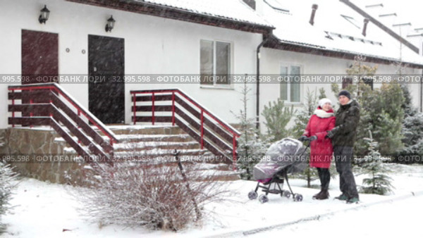 Happy couple discussing new townhouse while walking with baby stroller. Family standing outdoor at strong snowfall. Стоковое видео, видеограф Кекяляйнен Андрей / Фотобанк Лори