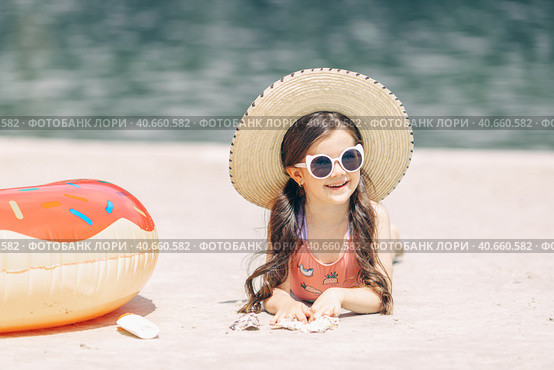 Happy cute smiling little girl in sunglasses lying on the sand on a beach in a swimsuit in a straw hat. Стоковое фото, фотограф Ирина Михайличенко / Фотобанк Лори