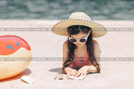 Happy cute smiling little girl in sunglasses lying on the sand on a beach in a swimsuit in a straw hat. Стоковое фото, фотограф Ирина Михайличенко / Фотобанк Лори