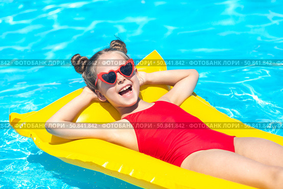 Happy laughing little girl with open mouth, in red swimsuit lies on yellow inflatable mattress in swimming pool with blue water on summer day on tropical vacation. Relaxation, fun, carefree childhood. Стоковое фото, фотограф Ирина Михайличенко / Фотобанк Лори
