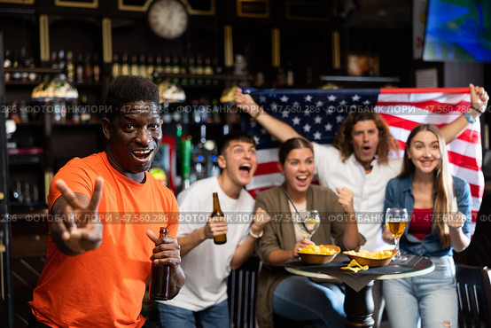 Happy multiracial sport fans holding the flag of USA, enjoing game, beer and chips in the bar. Стоковое фото, фотограф Яков Филимонов / Фотобанк Лори