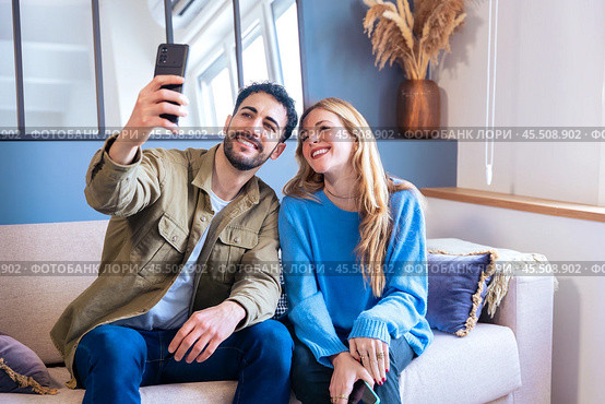 Happy young couple takes a selfie together with smartphone on sofa at home. Стоковое фото, фотограф Raquel Corcoles Mendez / Фотобанк Лори